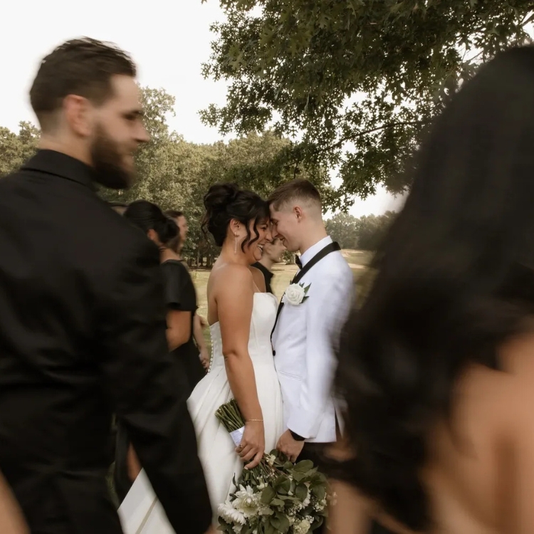 Bride and groom standing close together with wedding party blurred in the foreground at Running Deer Golf Club